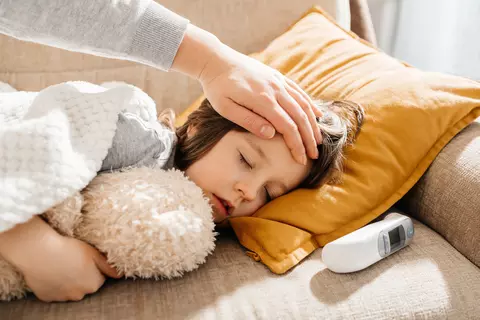Nemocné dítě spí na gauči, matka rukou kontroluje teplotu na čele