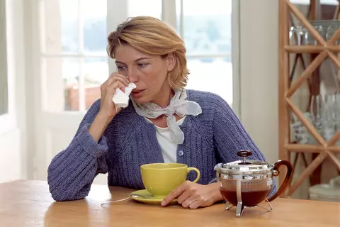 Žena se známkami nachlazení si utírá nos, je doma, pije čaj a kolem krku má šátek