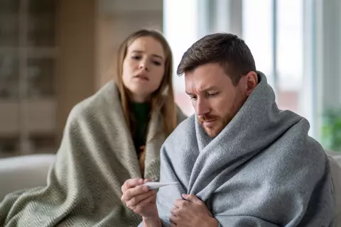 Mladý pár je doma na gauči, muž drží teploměr a oba jsou zabaleni do deky, vypadají nemocně
